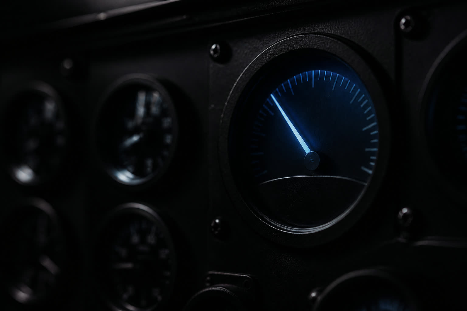 A dark instrument panel with one illuminated needle and unused gauges in shadow